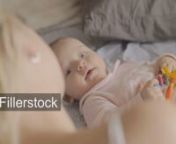 A cute baby girl in a pink romper suit is lying on a back on a bed and holding a colorful toy in hands. She is smiling to her mother, who is right next to her, gently holding a hand on babys bellynLicense this clip: https://fillerstock.com/video/15580